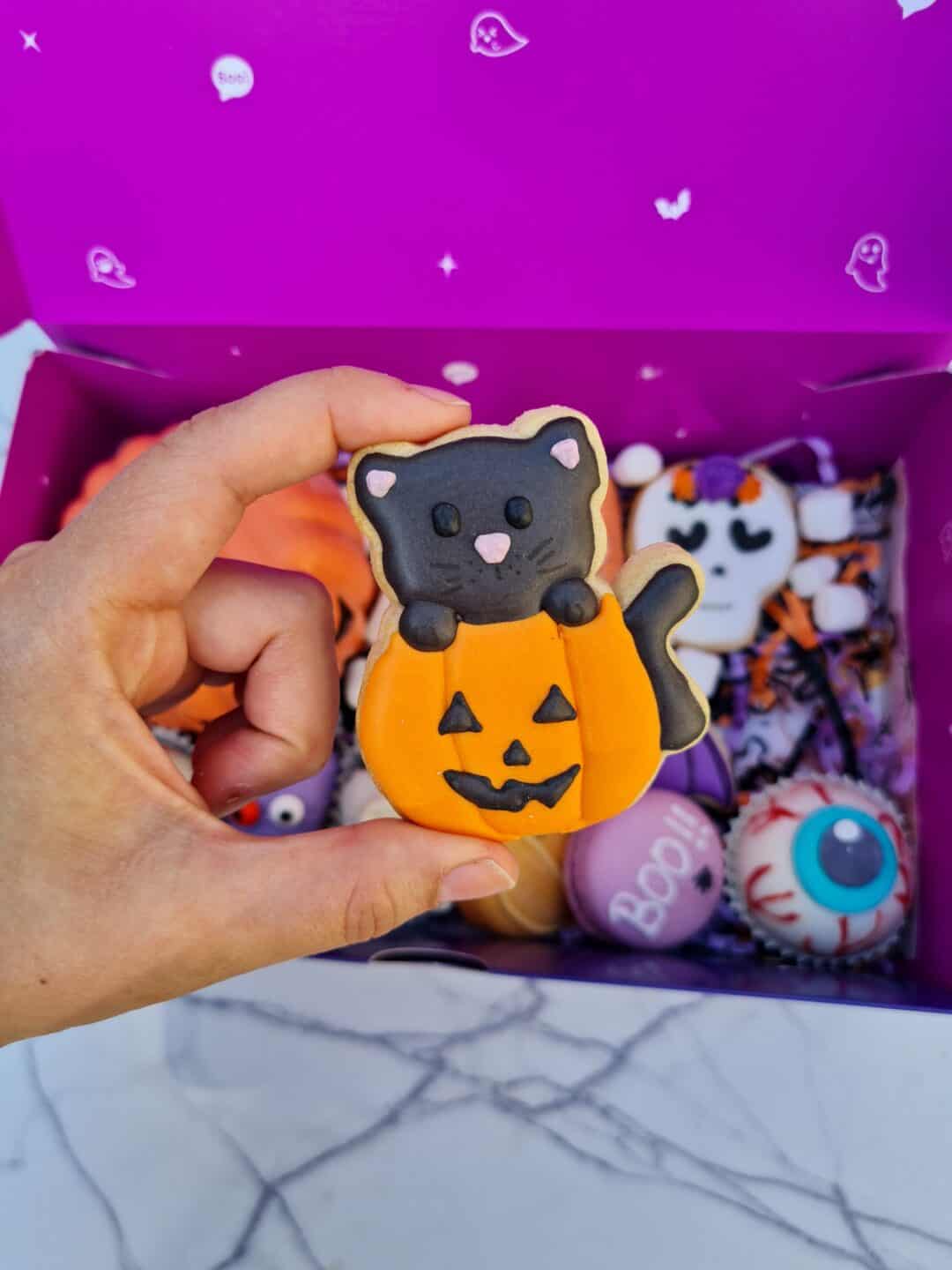 Galleta decorativa de gato con calabaza para Halloween, en caja con otros dulces temáticos.